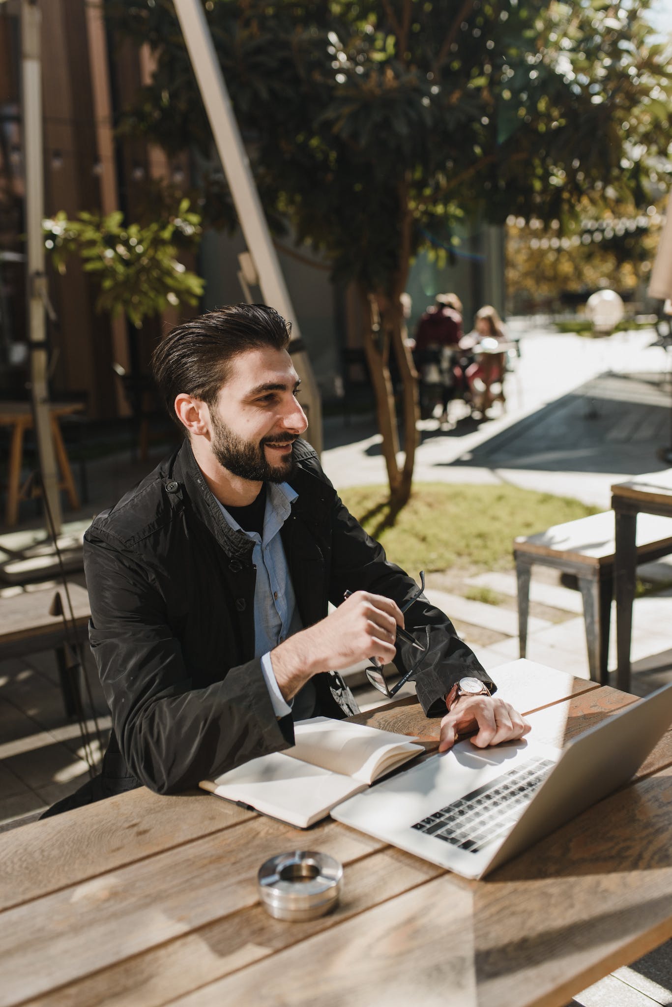 Smiling ethnic entrepreneur in trendy clothes sitting at wooden table with notebook and browsing laptop while working on street in sunny day